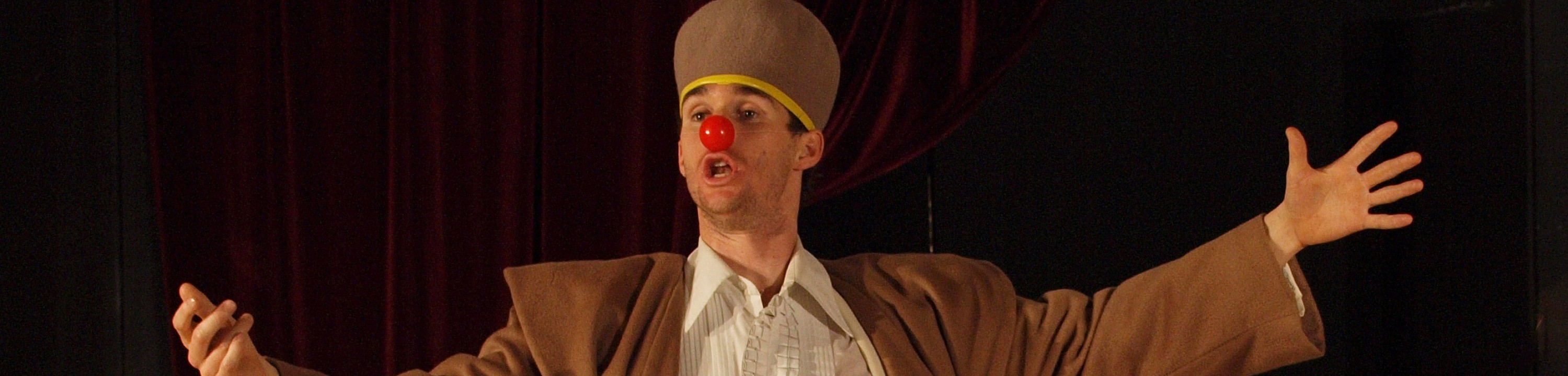a clown with a red nose gesturing on stage - image credit Helikos